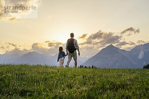 Father and daughter hiking in meadow by mountains at sunset