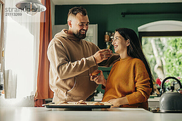 Smiling man feeding cinnamon bun to pregnant wife in kitchen at home