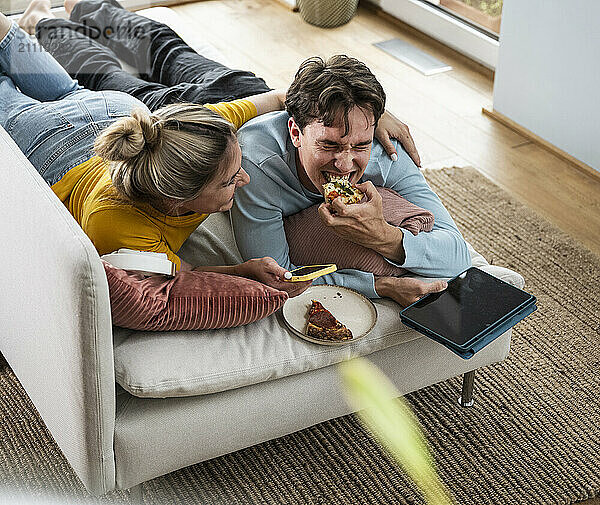 Smiling woman with arm around boyfriend eating pizza lying on sofa at home