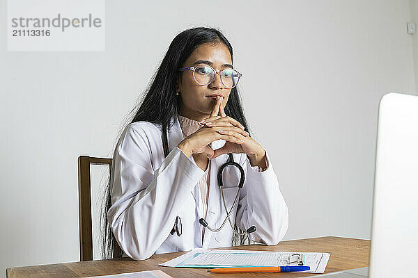 Young doctor in lab coat sitting with reports on table at clinic