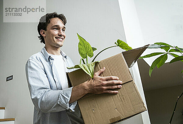 Smiling young man carrying cardboard box with houseplant near wall