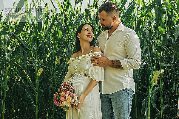 Pregnant woman holding bouquet of flowers and standing with husband near corn field