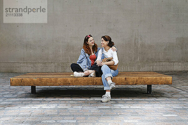 Happy lesbian couple sitting on bench near wall at sidewalk