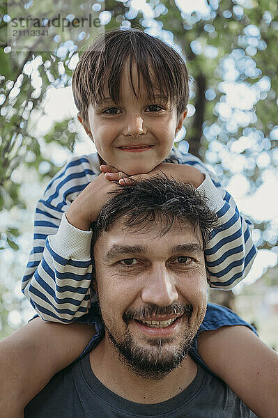 Smiling father carrying son on shoulders at tree