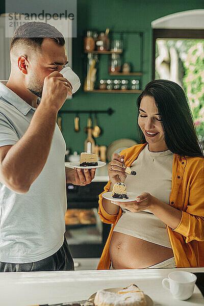 Husband with pregnant wife drinking tea and eating cake in kitchen