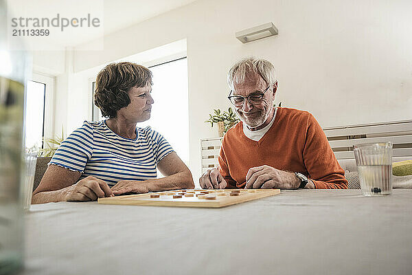 Smiling man sitting near table and playing board game with wife at home