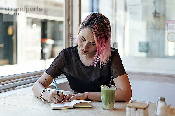 Woman with pink hair writing in diary at cafe