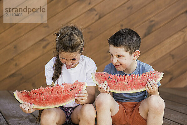 Playful siblings with watermelon slices making faces and sitting at porch