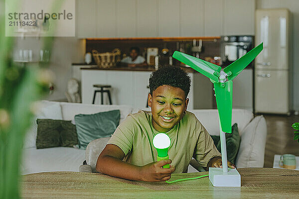 Smiling boy powering light bulb through wind turbine model at home