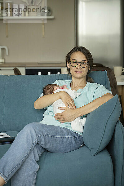 Smiling mother carrying baby in arms and sitting on sofa at home