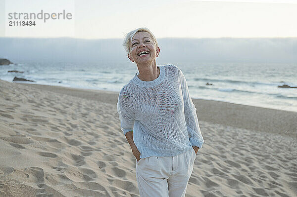 Cheerful mature woman with hands in pockets enjoying leisure time at beach