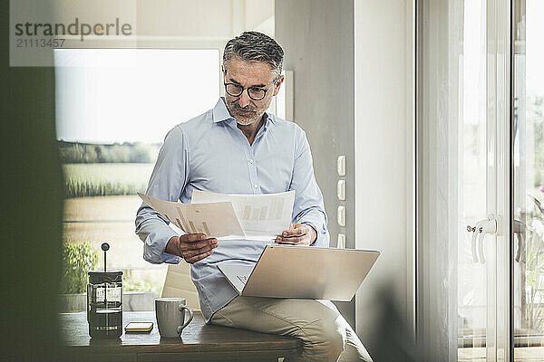 Businessman examining documents sitting with laptop at home office