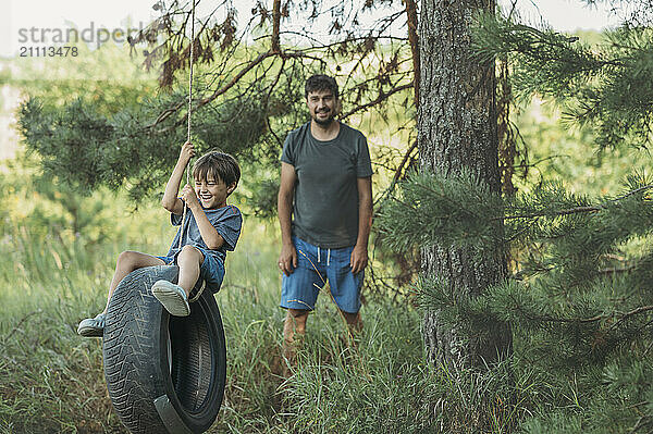 Boy having fun on tire swing near father at forest