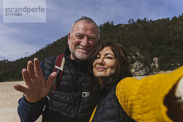 Smiling woman taking selfie with man waving at beach