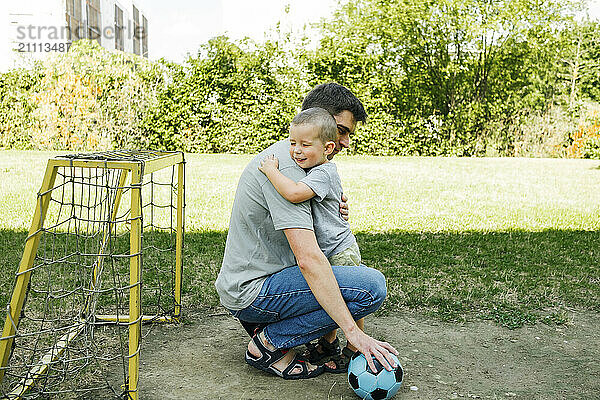 Son embracing father crouching near soccer net in playground