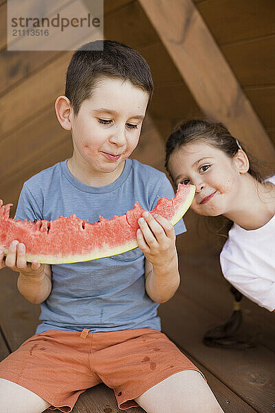 Brother looking at watermelon slice and sitting with sister outside house