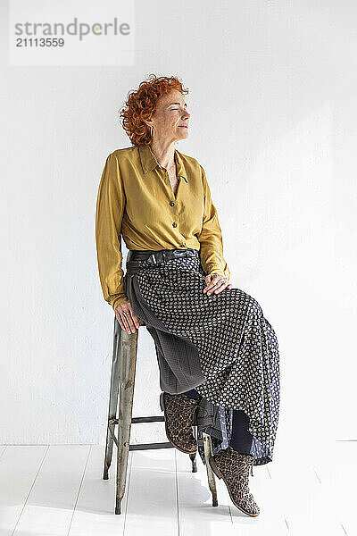 Senior woman with eyes closed sitting on stool in front of white wall