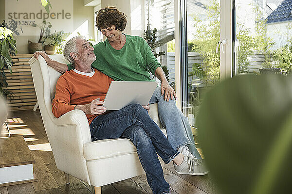 Happy senior couple with laptop sitting on chair at home