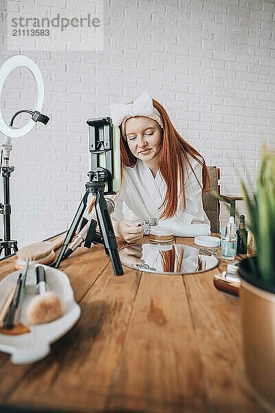 Teenager girl creating beauty vlog tutorial with makeup products on a wooden table in a modern indoor setting.