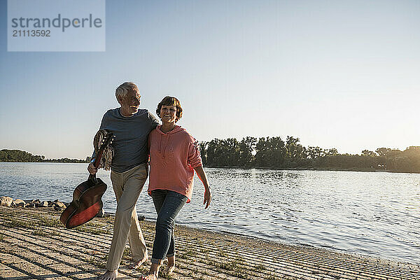 Happy senior man holding guitar and walking with woman near river