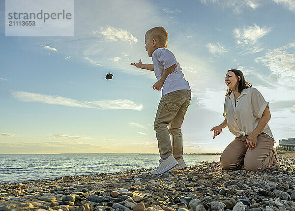 The boy throws a stone into the water on the beach with his mother.