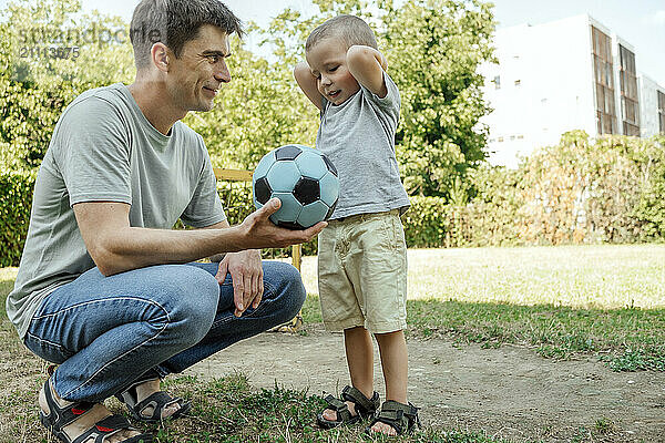 Father holding soccer ball crouching near son in playground