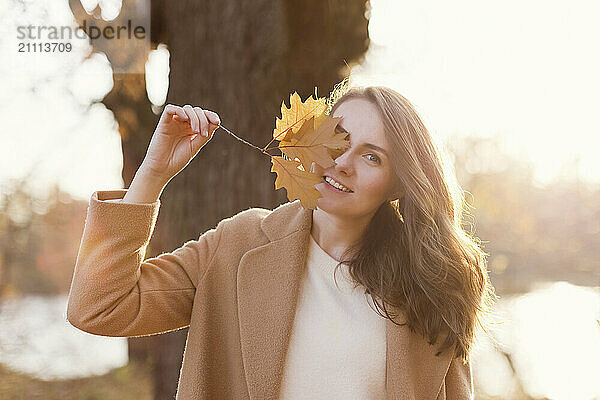 Smiling woman wearing beige coat covering eye with autumn leaf at forest