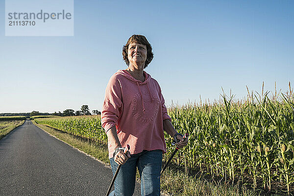 Smiling senior woman holding hiking poles and walking on road near agricultural field