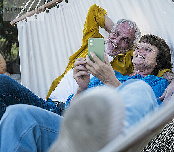 Affectionate senior man lying on hammock with woman using smart phone at back yard