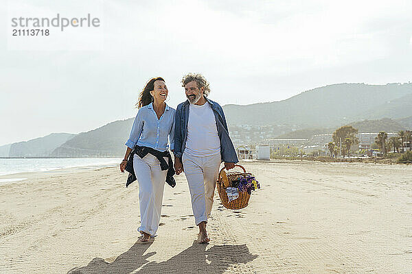 Happy couple holding hands and walking with basket at beach on sunny day