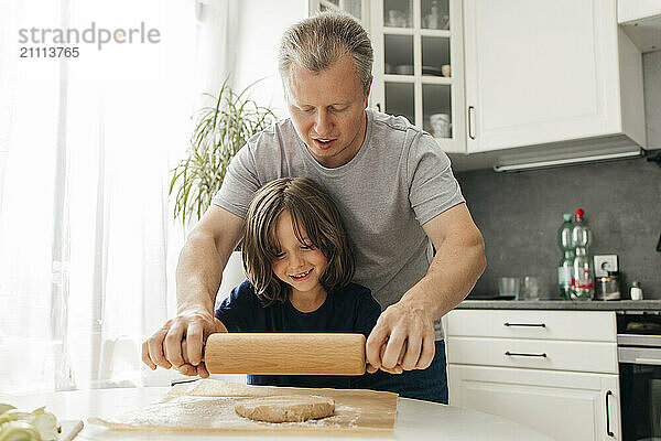 Father helping son make dough using rolling pin in kitchen