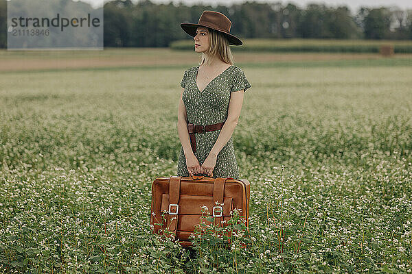 Beautiful woman holding suitcase and standing amidst field