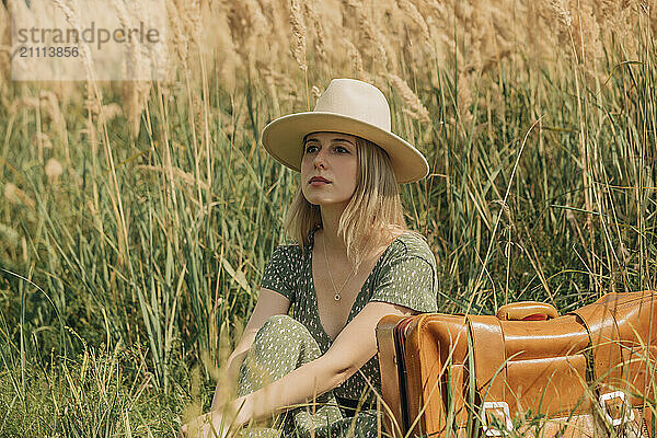 Thoughtful woman with hat sitting amidst reeds on sunny day