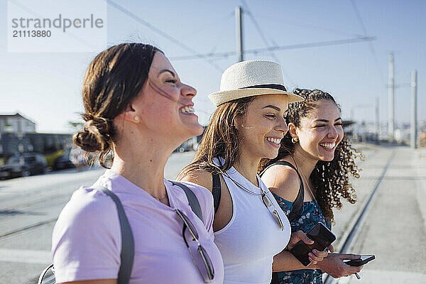 Happy friends with smart phones standing at tram tracks on sunny day