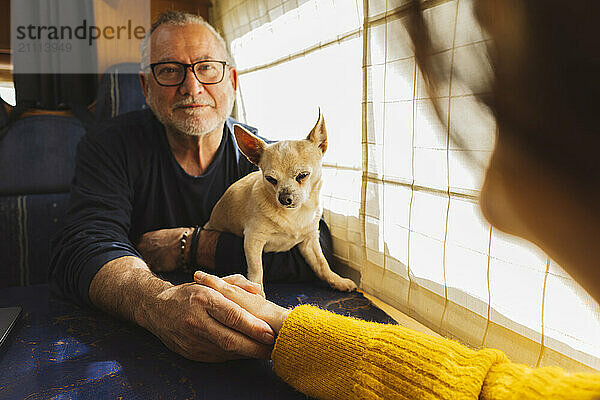 Senior man sitting with Chihuahua dog and holding woman's hand in motor home