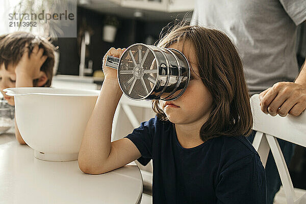 Boy holding flour sifter and sitting near table in kitchen