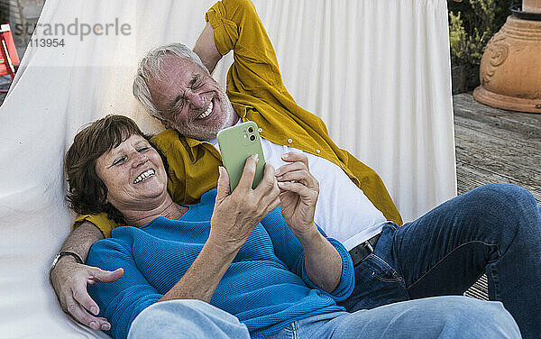 Happy senior woman using smart phone and lying on hammock with man at back yard