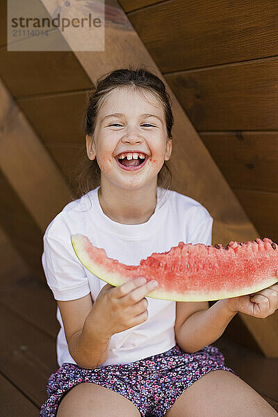 Laughing girl holding slice of watermelon and sitting at porch