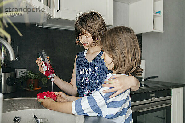 Affectionate girl pouring dishwashing liquid and helping brother in kitchen