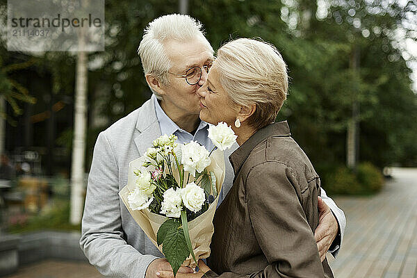 Senior man kissing woman with bouquet at street