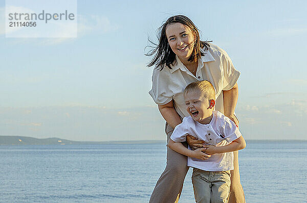 A dark-haired happy woman in a white shirt with her son on the beach.