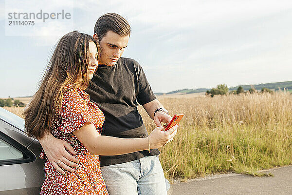 Affectionate couple standing and using smart phone near car