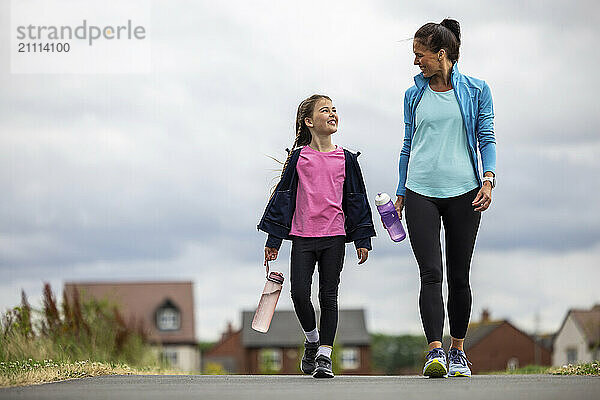 Mother and daughter holding water bottles and walking on footpath