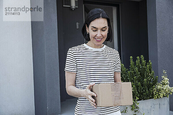 Happy woman holding box and standing in front of house