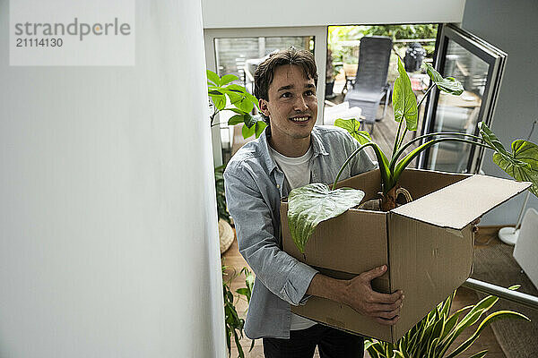 Young man carrying cardboard box with houseplant at new apartment