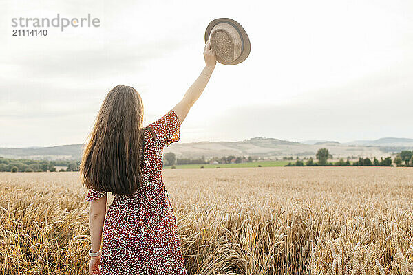 Young woman holding hat with hand raised in spikelet field