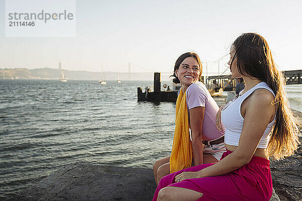 Smiling friends sitting together on promenade near water