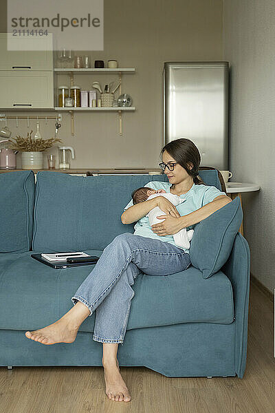 Mother carrying baby in arms and sitting on sofa at home