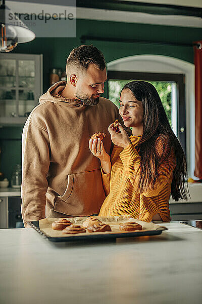 Young husband looking at pregnant wife enjoying cinnamon bun in kitchen