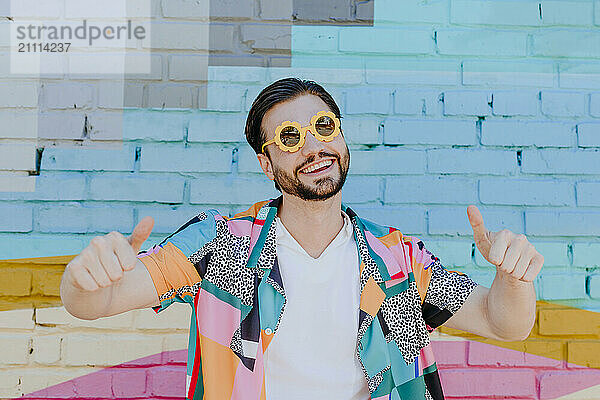 Happy man wearing novelty glasses and gesturing thumbs up in front of brick wall